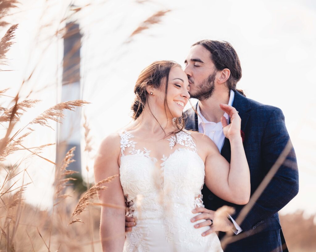 Bride and groom share a sweet moment on a beach with a lighthouse behind them.
